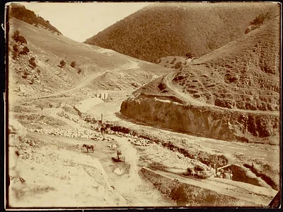 View of river bend with group of people on right, horse or burro mid-ground, wooded hill in background