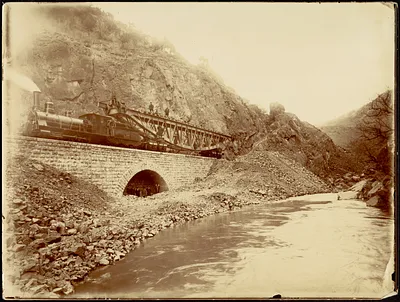Section of steel bridge being transported by train across stone viaduct, men standing under arch
